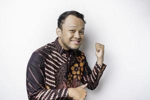 Excited Asian man wearing batik shirt pointing at the copy space beside him, isolated by white background photo