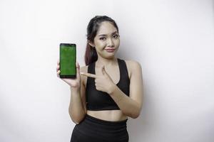 A thoughtful young sporty woman is wearing sportswear while holding her chin and showing green screen on her phone, isolated by white background photo