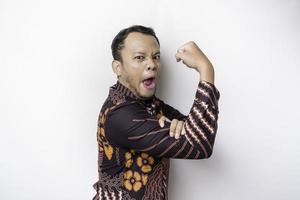 Excited Asian man wearing batik shirt showing strong gesture by lifting his arms and muscles smiling proudly photo