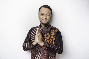 Smiling young Asian man wearing batik shirt, gesturing traditional greeting isolated over white background photo