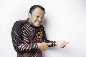 Excited Asian man wearing batik shirt pointing at the copy space beside him, isolated by white background photo