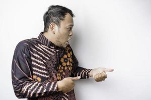 Shocked Asian man wearing batik shirt pointing at the copy space beside him, isolated by white background photo