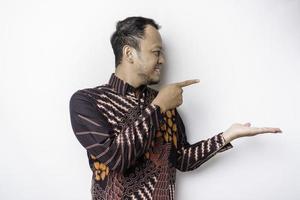Excited Asian man wearing batik shirt pointing at the copy space beside him, isolated by white background photo