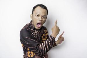 Serious Asian man wearing batik shirt pointing at the copy space on top of him, isolated by white background photo