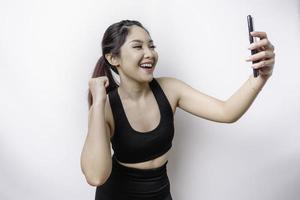A young sporty Asian woman with a happy successful expression wearing sportswear and holding smartphone isolated by white background photo