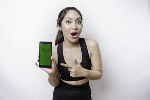 Shocked sporty Asian woman wearing sportswear and showing green screen on her phone, isolated by white background photo