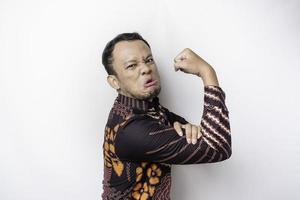 Excited Asian man wearing batik shirt showing strong gesture by lifting his arms and muscles smiling proudly photo