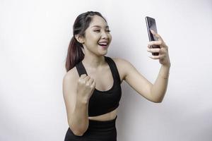 A young sporty Asian woman with a happy successful expression wearing sportswear and holding smartphone isolated by white background photo