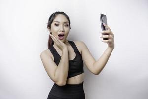 sorprendida mujer asiática deportiva usando ropa deportiva y sosteniendo su teléfono, aislada por fondo blanco foto