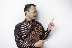 Excited Asian man wearing batik shirt pointing at the copy space beside him, isolated by white background photo