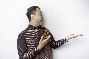 Shocked Asian man wearing batik shirt pointing at the copy space beside him, isolated by white background photo