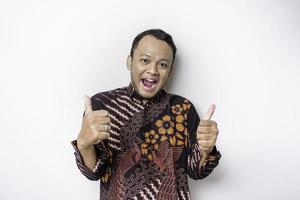 Excited Asian man wears batik shirt, gives thumbs up hand gesture of approval, isolated by white background photo