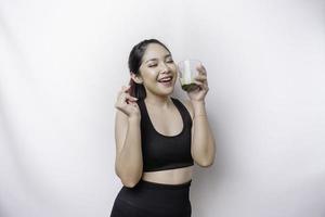 Joyful sporty Asian woman wearing sportswear with glass of tasty green smoothie, isolated on white background. photo
