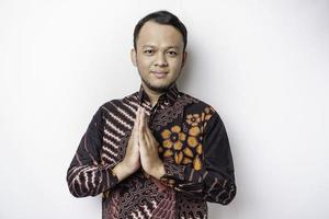 Smiling young Asian man wearing batik shirt, gesturing traditional greeting isolated over white background photo