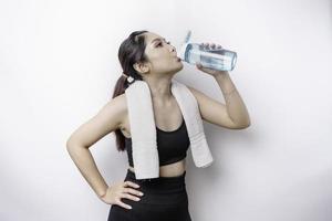 mujer asiática deportiva posando con una toalla en el hombro y bebiendo de una botella de agua, sonriendo y relajándose después del entrenamiento foto