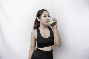 Joyful sporty Asian woman wearing sportswear is drinking a glass of tasty green smoothie, isolated on white background. photo