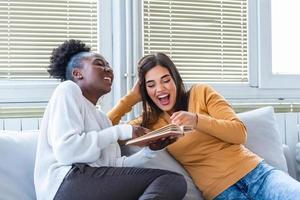 chicas divirtiéndose en casa, riendo y leyendo un libro divertido. diferentes amigos de raza en casa leyendo un libro y disfrutando de su tiempo juntos foto