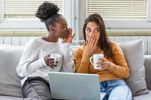 Diverse female friends sharing secrets. Two women gossiping at home. Excited emotional girl whispering to her friend ear while sitting in living-room photo