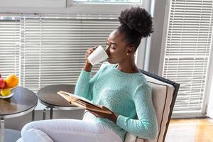 Young beautiful African American girl reading a book on the couch with the library bookshelves in the back. Beautiful woman on a white sofa reading a book photo