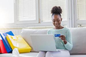 mujer joven en el sofá de compras en línea con la computadora portátil. joven negra ingresando información de la tarjeta mientras compra en línea. concepto de compras en línea foto