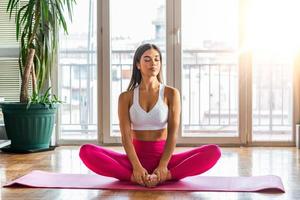Portrait of beautiful young woman working out against big windows, doing yoga or pilates exercise, sitting in baddha konasana, bound angle, cobbler, butterfly pose. Full length shot photo