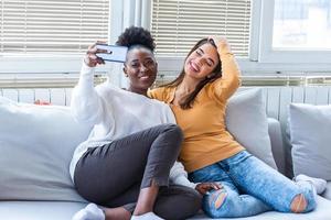 Two beautiful young women take selfie on sunny day at home. Girlfriends. Communication and gossip. Lovable caucasian and African American girls expressing positive emotions to camera. photo