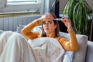 Sickness, seasonal virus problem concept. Woman being sick having flu lying on sofa looking at temperature on thermometer. Sick woman lying in bed with high fever. Cold flu and migraine. photo