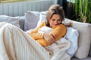 Woman in painful expression holding hands against belly suffering menstrual period pain, lying sad on home bed, having tummy cramp in female health concept photo