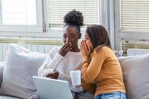 Diverse female friends sharing secrets. Two women gossiping at home. Excited emotional girl whispering to her friend ear while sitting in living-room photo