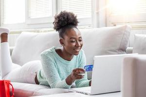 mujer joven en el sofá de compras en línea con tarjeta de débito. hermosa chica negra que usa una computadora portátil para comprar en línea en casa foto