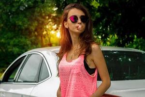 glamour brunette in dark glasses with chewing gum in the mouth stands near the car photo