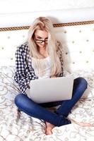 Photo of beauty young blonde woman with blue eyes sits on bed with laptop