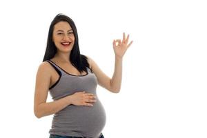 happy young brunette pregnant girl with hand on her belly showing OK isolated on white background photo