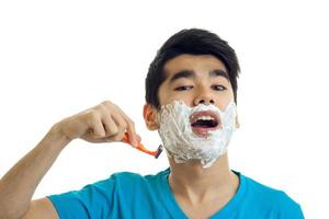 young cute guy with foam on his face opened his mouth and shaves his beard machine close-up