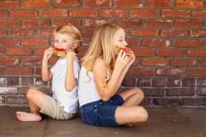 Cute Young Cuacasian Boy and Girl Eating Watermelon Against Brick Wall photo