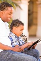 African American Father and Mixed Race Son Using Computer Tablet on Bench in Park photo