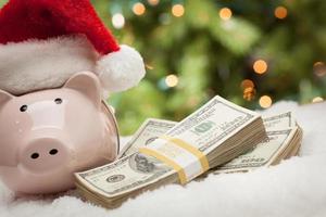 Piggy Bank Wearing Santa Hat Near Stacks of Hundred Dollar Bills on Snowflakes photo