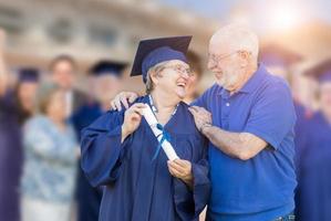 Senior Adult Woman In Cap and Gown Being Congratulated By Husband At Outdoor Graduation Ceremony. photo