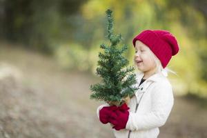 niña en guantes rojos y gorra sosteniendo un pequeño árbol de navidad
