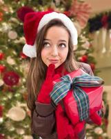 Thinking Girl Wearing A Christmas Santa Hat with Bow Wrapped Gift In Front of Decorated Tree photo