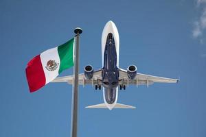 Vista inferior del avión de pasajeros que sobrevuela ondeando la bandera de México en el poste foto