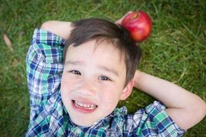 Mixed Race Chinese and Caucasian Young Boy With Apple Relaxing On His Back Outside On The Grass. photo