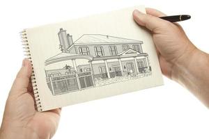 manos sosteniendo bolígrafo y bloc de papel con dibujo de casa