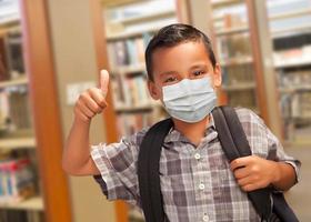 estudiante hispano con mascarilla con los pulgares hacia arriba y mochila en la biblioteca foto