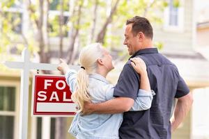 Pareja caucásica mirando y apuntando al frente de la casa y el cartel de venta de bienes raíces foto