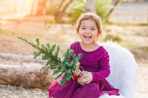 Linda niña de raza mixta sosteniendo un pequeño árbol de navidad al aire libre foto