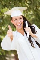 Niña de raza mixta Thumbs up celebrando la graduación afuera con toga y birrete con diploma en la mano foto