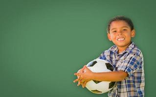 Cute Young Mixed Race Boy Holding Soccer Ball In Front of Blank Chalk Board photo