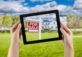 manos femeninas sosteniendo la tableta de la computadora con el cartel de venta de casa en la pantalla, detrás de la casa. foto