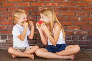 Cute Young Cuacasian Boy and Girl Eating Watermelon Against Brick Wall photo